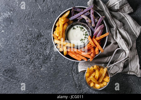 Variety of french fries traditional potatoes, purple potato, carrot served with white cheese sauce, salt, thyme in black bowl over dark texture backgr Stock Photo