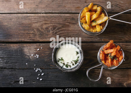 Variety of french fries traditional potatoes, sweet potato, carrot served in frying basket with white cheese sauce, salt, thyme over old wooden backgr Stock Photo