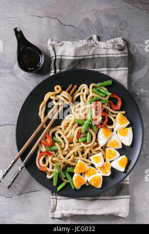 Cooking stir fry udon noodles, green beans, sliced paprika, boiled eggs, soy sauce with sesame seeds in black plate with wood chopsticks over gray tex Stock Photo