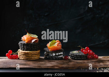 Stack of black charcoal and traditional crackers with smoked salmon, cream cheese, green salad and red currant berries on ood serving board over black Stock Photo