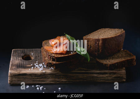 Stack of sliced homemade rye bread with smoked salmon, sea salt and fresh basil on wooden chopping board over dark black background. Stock Photo