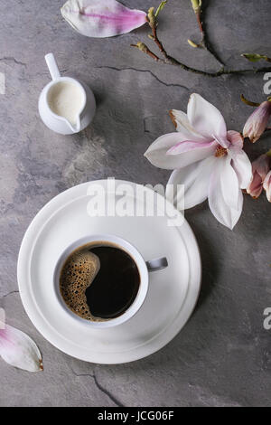 White cup of black coffee, served on white saucer with jug of cream and magnolia flower blossom branch over gray texture background. Flat lay, space Stock Photo