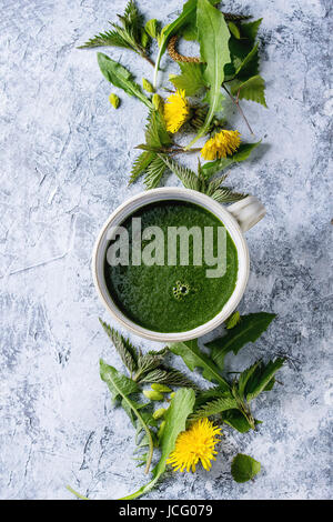 Spring green nettle and dandelion smoothie bowl served with yellow flowers, young birch leaves, spruce needles over gray blue texture background. Top Stock Photo