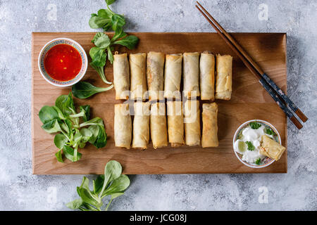 Fried spring rolls with red and white sauces, served on wood serving board with fresh green salad and wooden chopsticks over gray blue texture backgro Stock Photo
