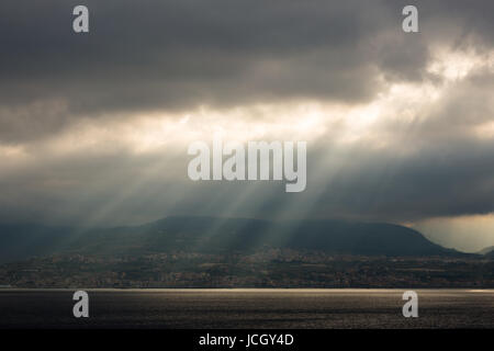 Landscape sunlight breaking through cloud viewed across the water from Messina port, Sicily looking towards Villa San Giovanni & Campo Calabro, Italy. Stock Photo