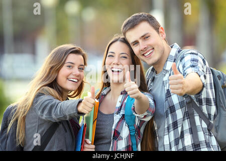 Three happy students looking at you with thumbs up in an university campus Stock Photo
