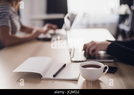 Close up picture of two businesswomen using tablet computer and laptop. Notebook with pen and cup of coffee in the foreground. Stock Photo