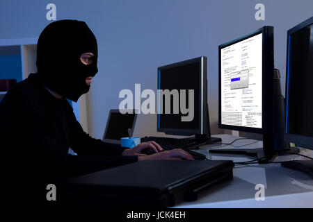 Masked hacker wearing a balaclava sitting at a desk downloading private information off a computer Stock Photo