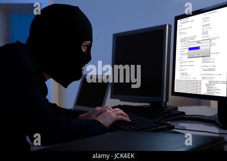 Masked hacker wearing a balaclava sitting at a desk downloading private information off a computer Stock Photo
