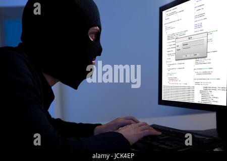 Masked hacker wearing a balaclava sitting at a desk downloading private information off a computer Stock Photo