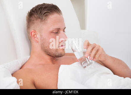 Man lying in bed propped up against the pillows drinking a glass of fresh water Stock Photo