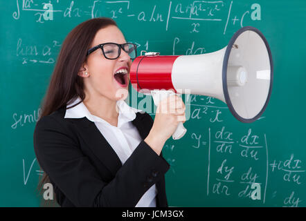 Angry young teacher shouting through megaphone against chalkboard in classroom Stock Photo