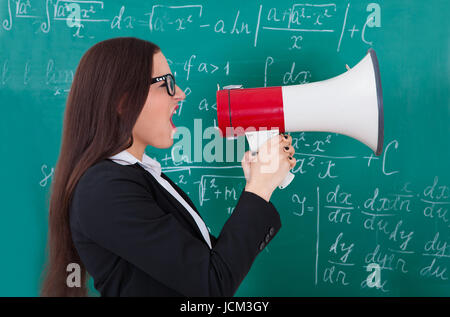 Angry young teacher shouting through megaphone against chalkboard in classroom Stock Photo