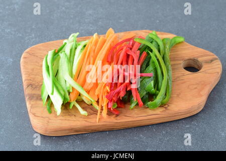 Fresh cucumber, carrot, red and green sweet paprika sliced in stripes on an olive wood cutting board on a grey abstract background. Step by step cooki Stock Photo