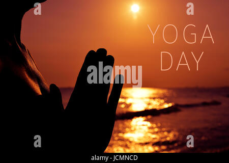 the silhouette of a young man with his hands put together as praying in front of the sea in backlight and the text yoga day Stock Photo