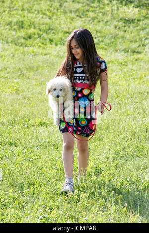 Pretty young girl carrying her little pet poodle under her arm as she walks across a grassy field in the sunshine looking down at it with a loving smi Stock Photo