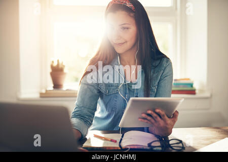 Cheerful young entrepreneur woman using laptop in earphones and holding tablet sitting in office. Stock Photo