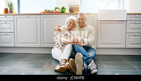 Happy mature attractive woman and handsome man embracing while sitting on kitchen floor. Large cabinets surround them. Stock Photo