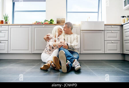 Cute senior couple looking at each other while seated comfortably on spacious dark tiled kitchen floor Stock Photo