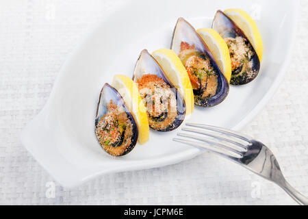 Grilled mussels cooked in the oven, accompanied by slices of lemon, grated pan and parsley. Mussels cooked on white plate. Stock Photo