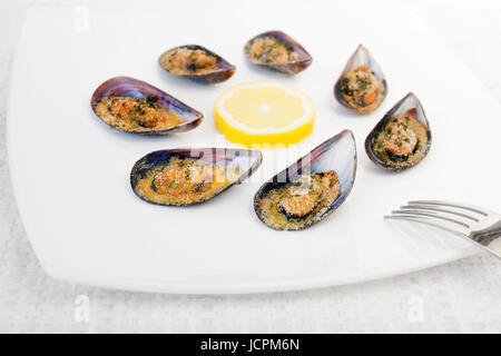 Grilled mussels cooked in the oven, accompanied by slices of lemon, grated pan and parsley. Mussels cooked on white plate. Stock Photo