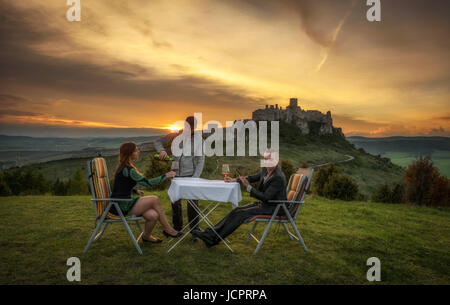 Party of three people drink red wine in nature under the ruins of a castle at sunset. Stock Photo