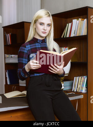 young girl student standing and reading book near bookshelves in library background Stock Photo