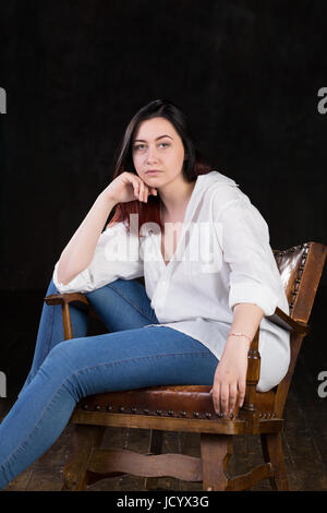 Beautiful white brown haired female with coloured in red ends, wearing men's white shirt and blue jeans, sitting in a vintage chair on a dark background Stock Photo