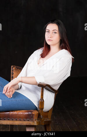 Beautiful white brown haired female with coloured in red ends, wearing men's white shirt and blue jeans, sitting in a vintage chair on a dark background Stock Photo