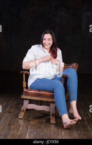 Beautiful white brown haired female with coloured in red ends, wearing men's white shirt and blue jeans, sitting in a vintage chair on a dark background Stock Photo