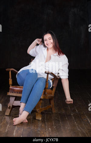 Beautiful white brown haired female with coloured in red ends, wearing men's white shirt and blue jeans, sitting in a vintage chair on a dark background Stock Photo