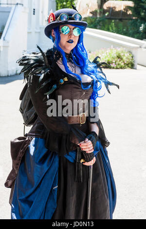 Steampunk Caucasian woman, 20s, large build, standing in sunshine with black costume and hat with goggles, long blue hair. Mirrored sunglasses, eye-contact. Stock Photo