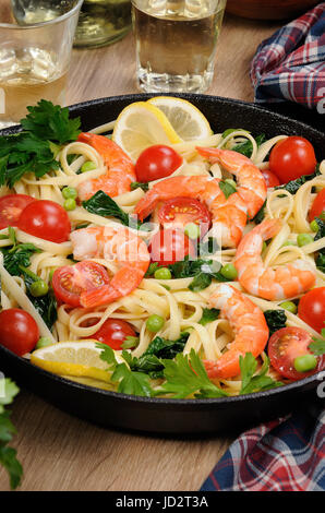 Pasta with fried prawns, peas, tomatoes and spinach in a frying pan, on a table with cider glasses. Stock Photo