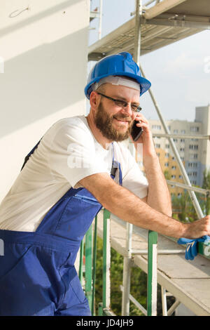 . Smiling construction worker in work outfit and helmet on head talking on the phone. Work at high altitude. Scaffolding in the background. Stock Photo