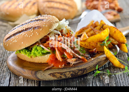Barbecue pulled pork sandwich with coleslaw, hot BBQ sauce and potato wedges in a paper bag served on a wooden table Stock Photo