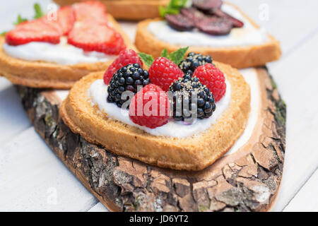 Heart shaped biscuits spread with quark, strawberries, blackberries, raspberries, cherries and a twig of mint presented on a tree disk