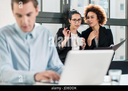 Two young women laughing while talking in the office behind their male colleague Stock Photo