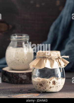 Active rye and wheat sourdough starter in glass jar on brown wooden background. Starter for sourdough bread. Toned image. Copy space. Vertical Stock Photo
