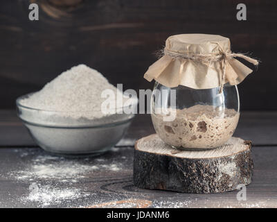 Active rye sourdough starter in glass jar and rye flour on brown wooden background. Starter for sourdough bread. Toned image. Copy space. Stock Photo