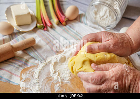 Old woman hands kneading dough for rhubarb pie and ingredients on the background Stock Photo