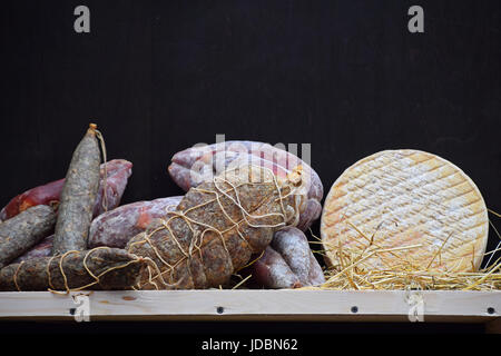 Wooden shelf with gourmet artisan cheese and sausage selection, on straw over black board background, close up, low angle view Stock Photo