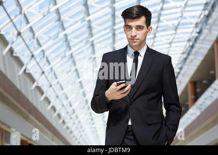 Portrait of confident successful businessman holding smartphone and posing, looking at camera in modern office building under glass roof Stock Photo