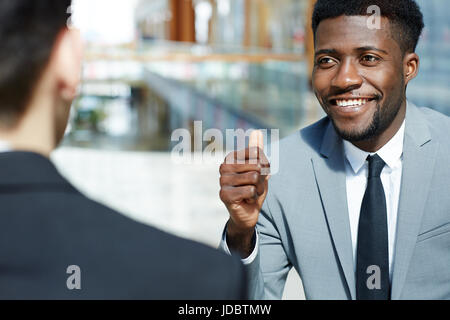 Portrait of young successful African-American businessman smiling and showing thumbs up while talking to partner, celebrating deal modern office build Stock Photo
