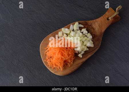 Grated carrots and cut in cubes onion on a olive wood cutting board on a grey abstract background. Step by step cooking Stock Photo