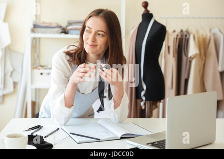 Smiling woman seamstress working with laptop and fabrics in workshop Stock Photo