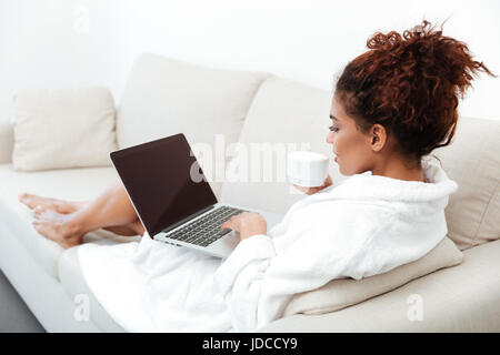 Back view picture of young african woman sitting on sofa at home and using laptop computer. Looking aside. Stock Photo