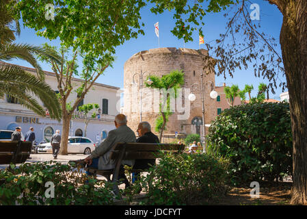 Old friends, rear view of two senior men chatting on a bench in public gardens near the medieval Torre di Porta Terra in Alghero Old Town, Sardinia Stock Photo