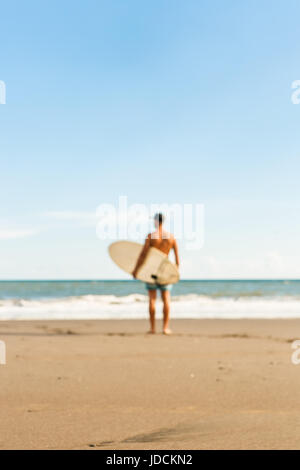 Out of focus, blurry image of handsome man stand with long surf surfing board wait for wave on surf spot at sea ocean beach. View from back. Concept o Stock Photo