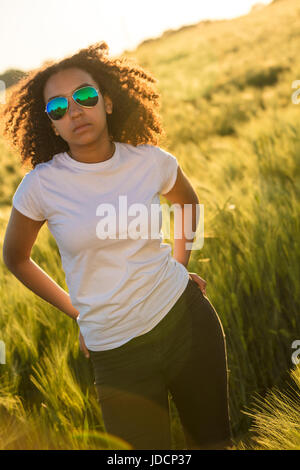 Beautiful mixed race African American female girl teenager young woman wearing reflective aviator sunglasses, jeans and a white t-shirt in a cornfield Stock Photo