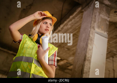 Beautiful young female construction architects at a construction site. She is holding plans and looking away. Stock Photo